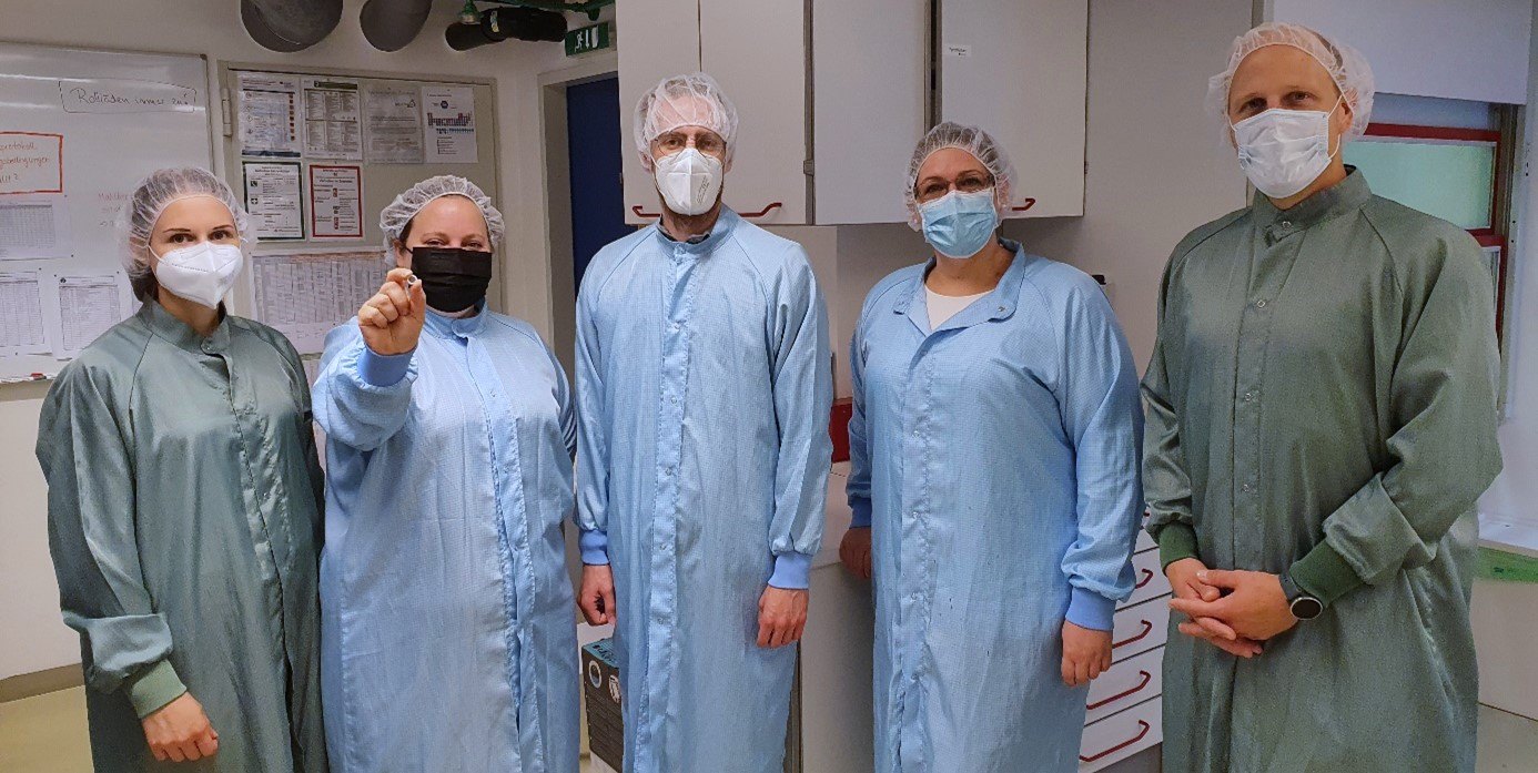 Eine Gruppe von Menschen in Schutzanzügen und mit Masken und Haarnetz posiert für die Kamera. Die zweite Person von Links hält eine Pulvertablette in die Kamera.